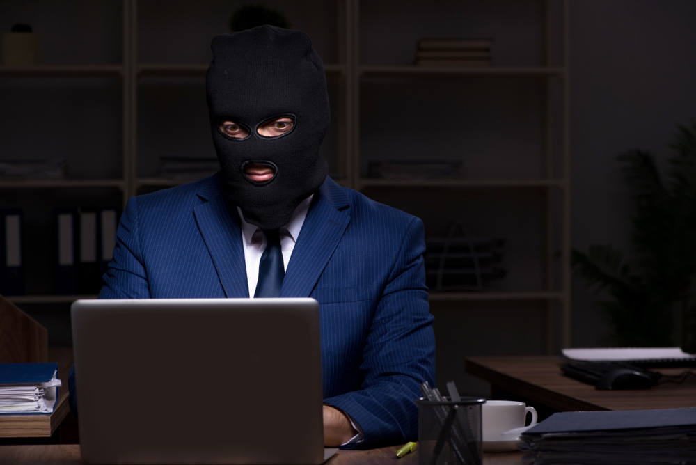 MaleEmployeeStealingInformationInTheOfficeNightTime - Tokenhell Person in a dark ski mask and blue suit sits at a desk typing on a laptop, suggesting a hacker.